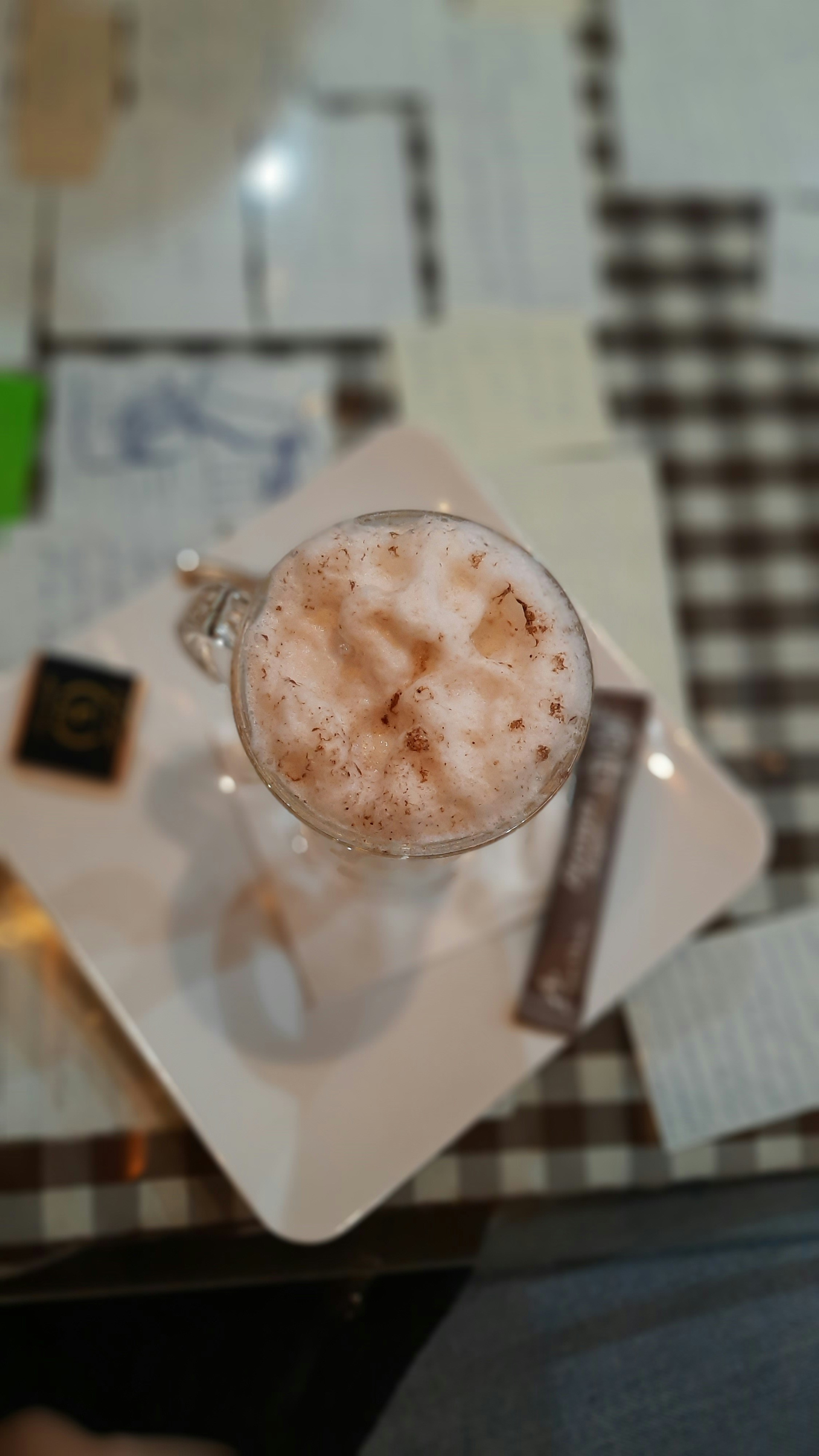 A frothy beverage topped with cinnamon is served in a glass mug on a white square plate. Next to the mug, there is a small dark chocolate square and a cocoa packet. The background reveals a checkered table covered in paper pieces.