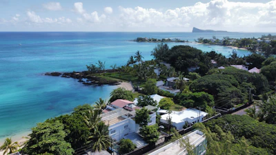 Aerial view of a tropical coastline with turquoise waters and greenery. There are several residential buildings with white rooftops nestled among lush trees. The shoreline is lined with palm trees, and there's a distant view of a larger island or landmass across the water.