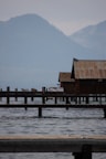 A serene lakeside scene featuring a rustic wooden building on stilts extending over the water. The background reveals misty, forested mountains under a cloudy sky. Several wooden piers stretch into the lake, and the water appears calm with gentle ripples.