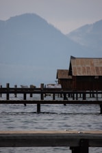 A serene lakeside scene featuring a rustic wooden building on stilts extending over the water. The background reveals misty, forested mountains under a cloudy sky. Several wooden piers stretch into the lake, and the water appears calm with gentle ripples.