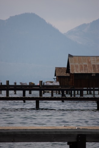 A serene lakeside scene featuring a rustic wooden building on stilts extending over the water. The background reveals misty, forested mountains under a cloudy sky. Several wooden piers stretch into the lake, and the water appears calm with gentle ripples.