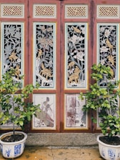 A decorative wooden panel featuring intricate carvings of animals and floral designs. The panel consists of four sections, each adorned with detailed artwork. In front, two potted plants with green foliage are placed symmetrically. The pots are white with blue artistic patterns.