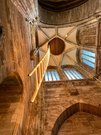 An intricately designed architectural interior with high, vaulted ceilings made of stone and wood. A striking bright yellow, illuminated ladder-like installation hangs vertically, adding a modern contrast to the historical setting. Tall arched windows with grid-patterned glass allow natural light to filter in.