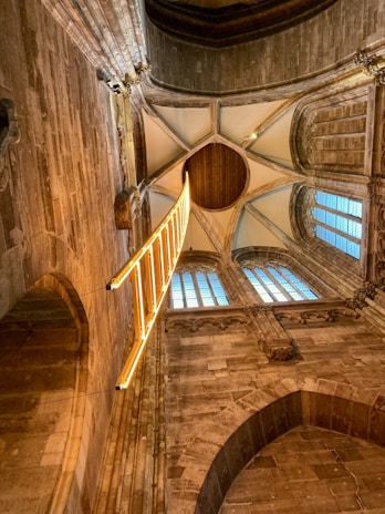 An intricately designed architectural interior with high, vaulted ceilings made of stone and wood. A striking bright yellow, illuminated ladder-like installation hangs vertically, adding a modern contrast to the historical setting. Tall arched windows with grid-patterned glass allow natural light to filter in.