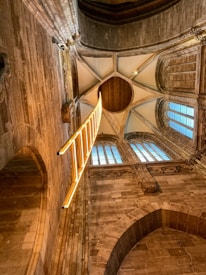 An intricately designed architectural interior with high, vaulted ceilings made of stone and wood. A striking bright yellow, illuminated ladder-like installation hangs vertically, adding a modern contrast to the historical setting. Tall arched windows with grid-patterned glass allow natural light to filter in.