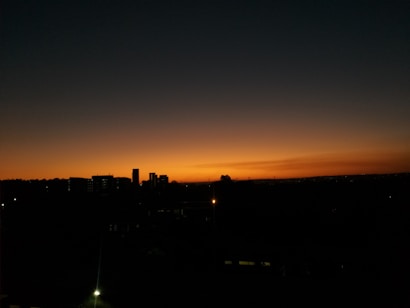 A serene dusk scene with a silhouetted cityscape against a vibrant sunset. The sky transitions from shades of dark blue to orange near the horizon. Buildings are visible as dark outlines, and a few lights are illuminated.