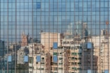 A grid-like pattern of window panes reflects a cityscape, capturing distorted images of modern high-rise buildings with varied architectural styles. The glass surface creates a mosaic of colors and shapes, adding an abstract quality to the urban reflection.