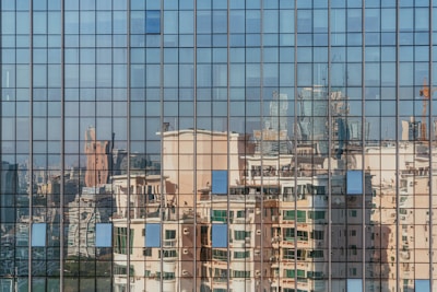 A grid-like pattern of window panes reflects a cityscape, capturing distorted images of modern high-rise buildings with varied architectural styles. The glass surface creates a mosaic of colors and shapes, adding an abstract quality to the urban reflection.