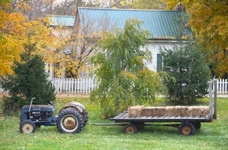 A rustic scene with a blue tractor hitched to a flatbed trailer loaded with hay bales. The trailer is parked on vibrant green grass in front of a white house with a green roof. The yard is framed by autumn-colored trees and a white picket fence.