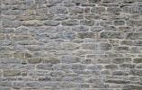 A textured stone wall composed of irregularly shaped stones in various sizes, tightly fitted together. The surface is rough and uneven, with subtle color variations and visible mortar lines between the stones.