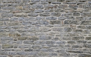 A textured stone wall composed of irregularly shaped stones in various sizes, tightly fitted together. The surface is rough and uneven, with subtle color variations and visible mortar lines between the stones.