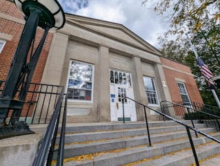 A front view of a United States Post Office building with brick and stone facade. The entrance is approached by a set of wide steps with metal railings. A flagpole with the American flag is on the right, and the sky is partly cloudy.