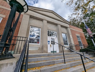A front view of a United States Post Office building with brick and stone facade. The entrance is approached by a set of wide steps with metal railings. A flagpole with the American flag is on the right, and the sky is partly cloudy.
