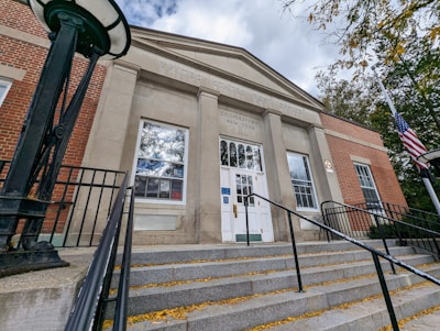 A front view of a United States Post Office building with brick and stone facade. The entrance is approached by a set of wide steps with metal railings. A flagpole with the American flag is on the right, and the sky is partly cloudy.