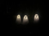 Three ghost decorations are illuminated in a dark setting. Each ghost is covered by a white sheet and has a different facial expression cut out of black material: one with a smiling face, one with an 'O' shaped mouth, and one with a sad face. Thin strands of dry grass are visible in the background.
