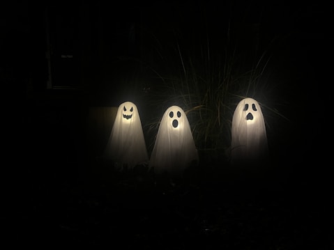 Three ghost decorations are illuminated in a dark setting. Each ghost is covered by a white sheet and has a different facial expression cut out of black material: one with a smiling face, one with an 'O' shaped mouth, and one with a sad face. Thin strands of dry grass are visible in the background.
