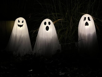 Three ghost decorations made of white sheets with black facial features stand illuminated in the dark, surrounded by tall grass. Each ghost has a unique face, with one smiling mischievously, one looking surprised, and the third appearing startled.