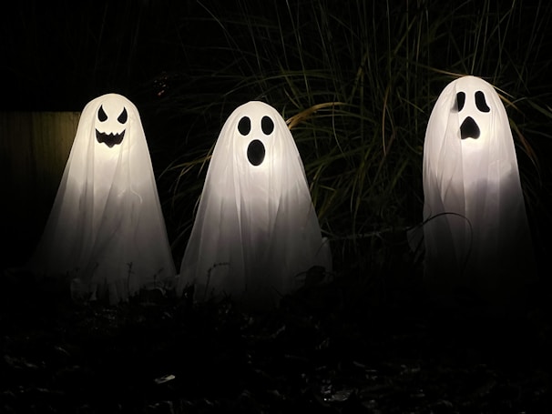 Three ghost decorations made of white sheets with black facial features stand illuminated in the dark, surrounded by tall grass. Each ghost has a unique face, with one smiling mischievously, one looking surprised, and the third appearing startled.