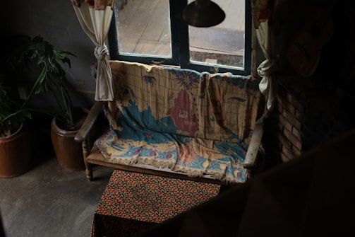 A cozy and rustic interior featuring a wooden bench covered with a colorful map-themed blanket. The room includes potted plants, a woven tablecloth with a floral pattern, and a window draped with tied-back curtains, filtering soft light into the space. A guitar rests against a brick wall, adding to the artistic and homely ambiance.