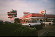 A vintage-style diner with a large, classic sign is prominently displayed. The diner has a retro design with a shiny chrome exterior and bright red accents. A classic car is parked in front, enhancing the nostalgic feel. The sky is partly cloudy, suggesting early evening or late afternoon lighting.