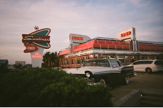 A vintage-style diner with a large, classic sign is prominently displayed. The diner has a retro design with a shiny chrome exterior and bright red accents. A classic car is parked in front, enhancing the nostalgic feel. The sky is partly cloudy, suggesting early evening or late afternoon lighting.
