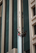 Two workers wearing helmets and harnesses are suspended on the side of a tall building. They are using ropes for support and carrying red bags, possibly for cleaning supplies. The building has a modern design with large reflective windows and alternating beige and gray vertical panels.