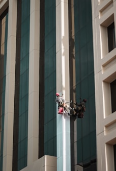 Two workers wearing helmets and harnesses are suspended on the side of a tall building. They are using ropes for support and carrying red bags, possibly for cleaning supplies. The building has a modern design with large reflective windows and alternating beige and gray vertical panels.