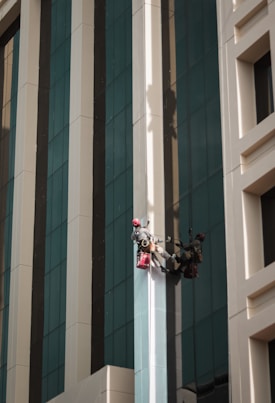 Two workers wearing helmets and harnesses are suspended on the side of a tall building. They are using ropes for support and carrying red bags, possibly for cleaning supplies. The building has a modern design with large reflective windows and alternating beige and gray vertical panels.