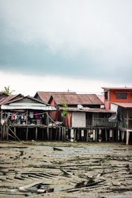 A series of stilt houses stand over a muddy, drying riverbed. The structures appear weathered and are constructed from a mix of materials like wood and metal. Clothes hang from a line outside one of the houses, adding a touch of color amid the muted tones. The sky is overcast, suggesting a gloomy or rainy atmosphere.
