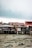 A series of stilt houses stand over a muddy, drying riverbed. The structures appear weathered and are constructed from a mix of materials like wood and metal. Clothes hang from a line outside one of the houses, adding a touch of color amid the muted tones. The sky is overcast, suggesting a gloomy or rainy atmosphere.