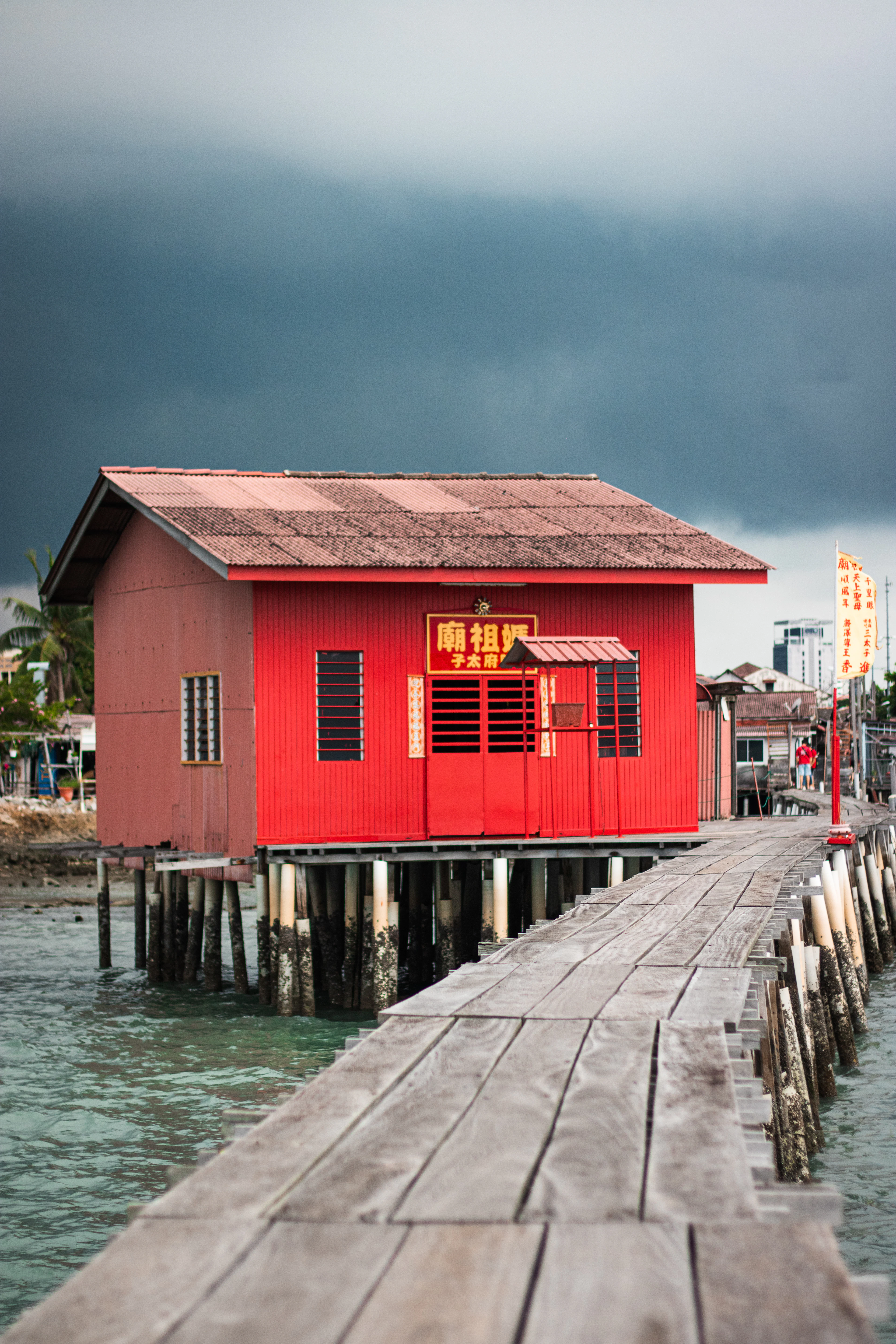 A red-painted timber shrine house raised on stilts beside the plank walkway of a clan jetty in George Town, Penang, its gold Chinese signboard glowing under a heavy slate storm sky.