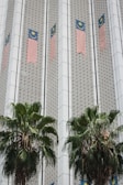 The image depicts a building facade featuring Malaysian flags aligned vertically along the structure. The facade has a patterned design with geometric elements, and two large palm trees are positioned in front of the building.