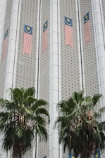 The image depicts a building facade featuring Malaysian flags aligned vertically along the structure. The facade has a patterned design with geometric elements, and two large palm trees are positioned in front of the building.