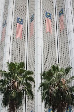 The image depicts a building facade featuring Malaysian flags aligned vertically along the structure. The facade has a patterned design with geometric elements, and two large palm trees are positioned in front of the building.