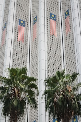 The image depicts a building facade featuring Malaysian flags aligned vertically along the structure. The facade has a patterned design with geometric elements, and two large palm trees are positioned in front of the building.
