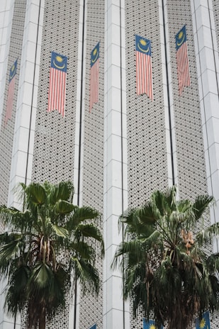 The image depicts a building facade featuring Malaysian flags aligned vertically along the structure. The facade has a patterned design with geometric elements, and two large palm trees are positioned in front of the building.
