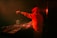 A person in a red hoodie is DJing, surrounded by electronic music equipment on a dimly lit stage. The background is dark, with hints of orange light creating a warm ambiance. The person is focused on mixing music, with one hand reaching out while operating the equipment.