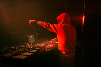 A person in a red hoodie is DJing, surrounded by electronic music equipment on a dimly lit stage. The background is dark, with hints of orange light creating a warm ambiance. The person is focused on mixing music, with one hand reaching out while operating the equipment.