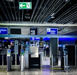 An airport security checkpoint with electronic gates and digital signage. Several signs display information for travelers, including an overhead green exit sign. The area is well-lit with modern design elements.