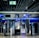 An airport security checkpoint with electronic gates and digital signage. Several signs display information for travelers, including an overhead green exit sign. The area is well-lit with modern design elements.