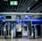 An airport security checkpoint with electronic gates and digital signage. Several signs display information for travelers, including an overhead green exit sign. The area is well-lit with modern design elements.