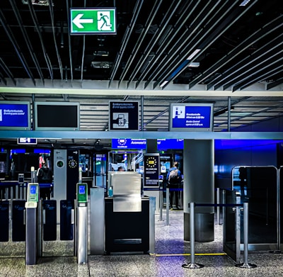 An airport security checkpoint with electronic gates and digital signage. Several signs display information for travelers, including an overhead green exit sign. The area is well-lit with modern design elements.