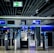 An airport security checkpoint with electronic gates and digital signage. Several signs display information for travelers, including an overhead green exit sign. The area is well-lit with modern design elements.