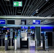 An airport security checkpoint with electronic gates and digital signage. Several signs display information for travelers, including an overhead green exit sign. The area is well-lit with modern design elements.