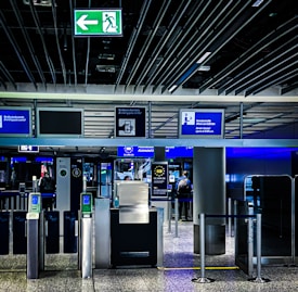 An airport security checkpoint with electronic gates and digital signage. Several signs display information for travelers, including an overhead green exit sign. The area is well-lit with modern design elements.
