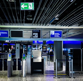 An airport security checkpoint with electronic gates and digital signage. Several signs display information for travelers, including an overhead green exit sign. The area is well-lit with modern design elements.