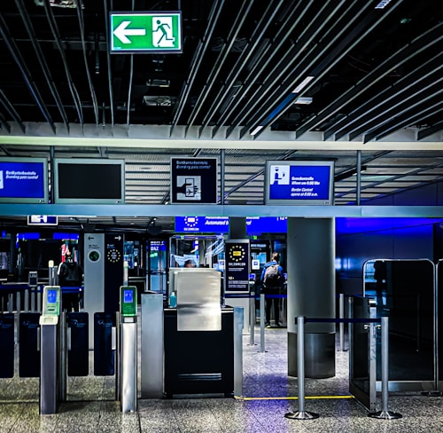An airport security checkpoint with electronic gates and digital signage. Several signs display information for travelers, including an overhead green exit sign. The area is well-lit with modern design elements.