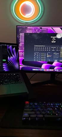 A workspace with two monitors displaying digital interfaces, one showing a weather application. A laptop is partially visible, and a mechanical keyboard is in the foreground. Neon circular wall art radiates vibrant colors above the monitors.