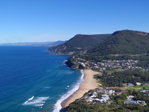 A coastal landscape with a sandy beach stretching along the shoreline, bordered by blue ocean waters and rolling waves. A green, forested hillside rises prominently in the background, with a small residential area nestled at the base of the hill. Houses and buildings are visible along the coastline, surrounded by trees and greenery.