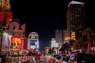 Bright and colorful Las Vegas cityscape at night with illuminated buildings and hotels. Neon signs and billboards light up the street. Cars are lined up on the road while palm trees are visible along the side.